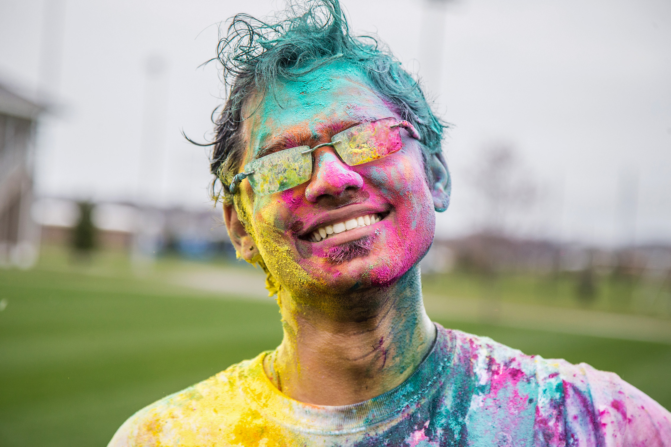 A person covered in colorful powder smiles broadly while wearing sunglasses, standing outdoors on a grassy field.