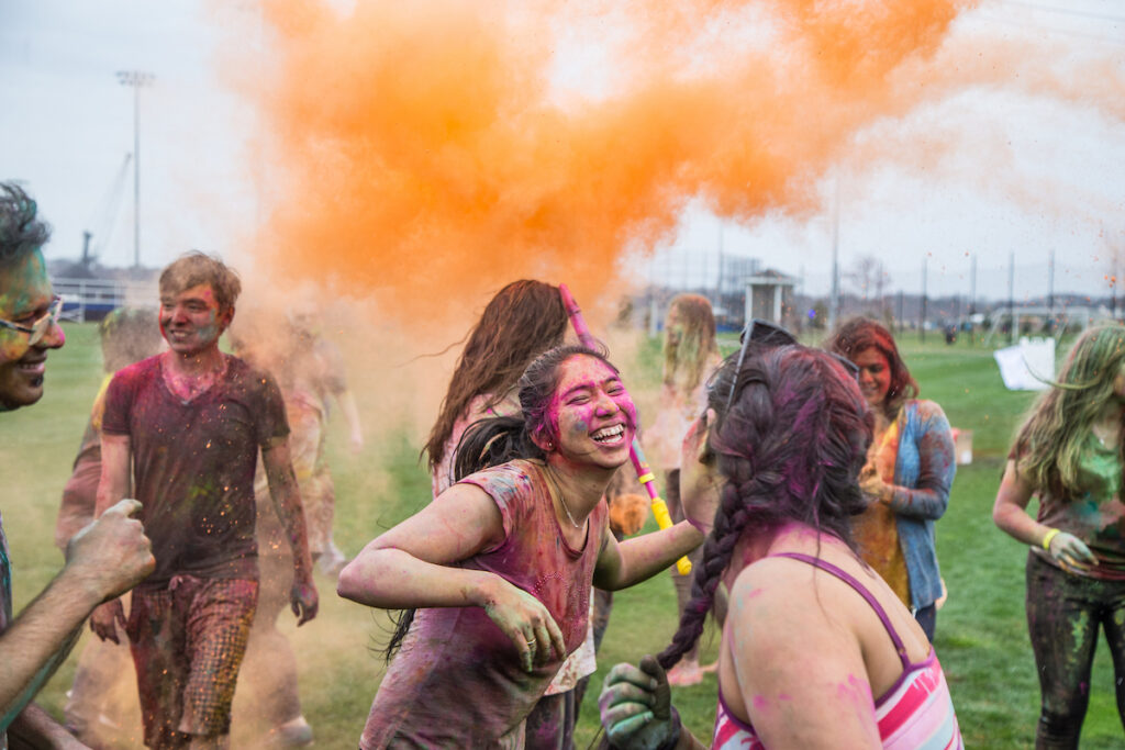 A group of people joyfully celebrate outdoors, covered in vibrant colored powder as a large orange cloud bursts overhead.