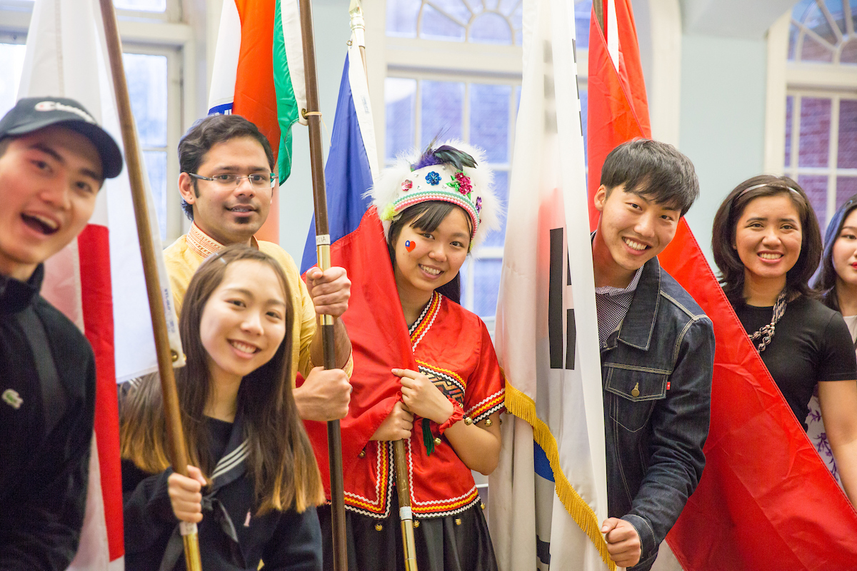 A group of diverse individuals smile while holding flags from various countries, including one person in traditional attire.