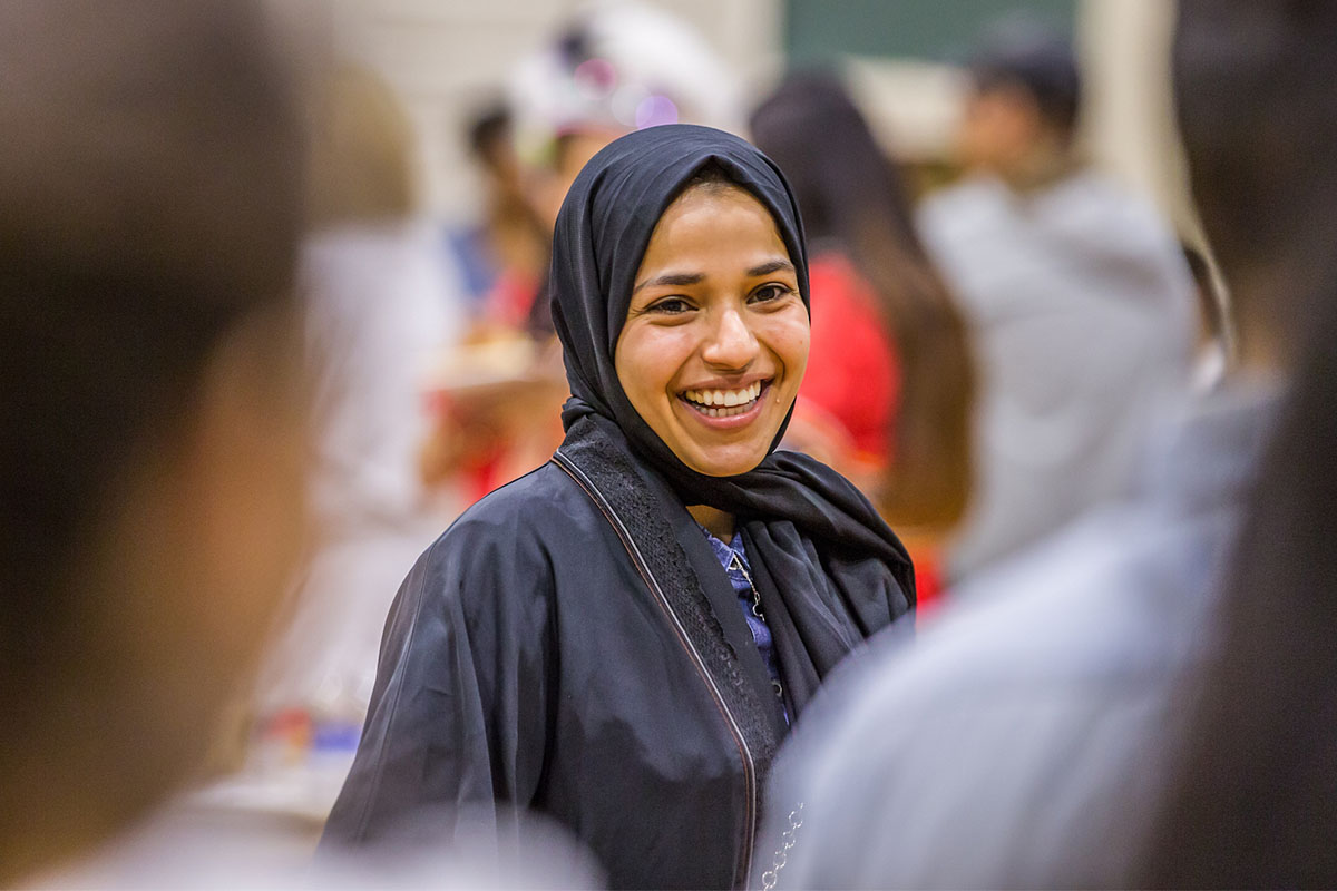 A person wearing a black hijab smiles warmly while engaging in conversation in a social gathering.