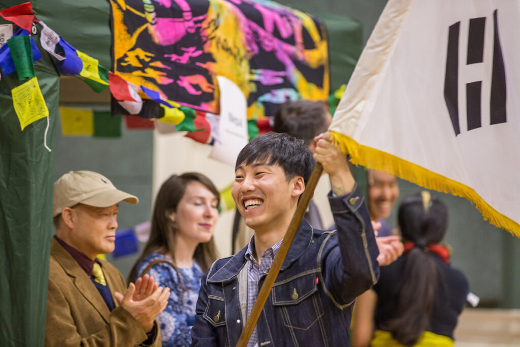 A smiling person in a denim jacket holds a flag with a black symbol, surrounded by others at an event adorned with colorful banners.