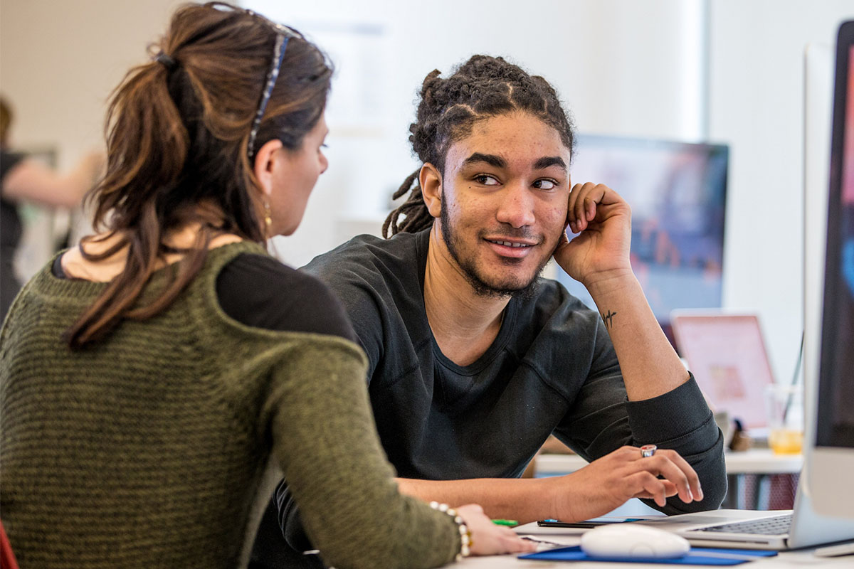 A student speaks with a professor at a desk with a computer.