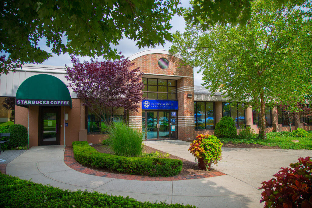 A brick building with a Starbucks coffee shop on the left and the entrance to Johnson & Wales University on the right, surrounded by greenery and trees.
