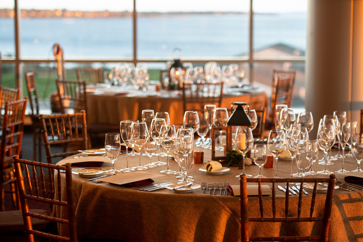 An elegantly set dining table with glassware and candles overlooks a waterfront view through large windows.