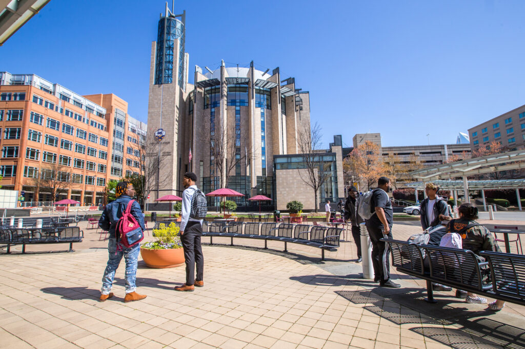 Students and pedestrians gather in a sunny plaza beside a modern architectural building with a prominent tower.