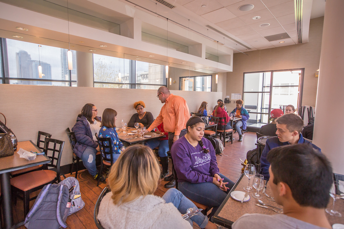 A group of people are seated at tables in a modern restaurant with large windows overlooking a city skyline, engaging in conversation and dining.