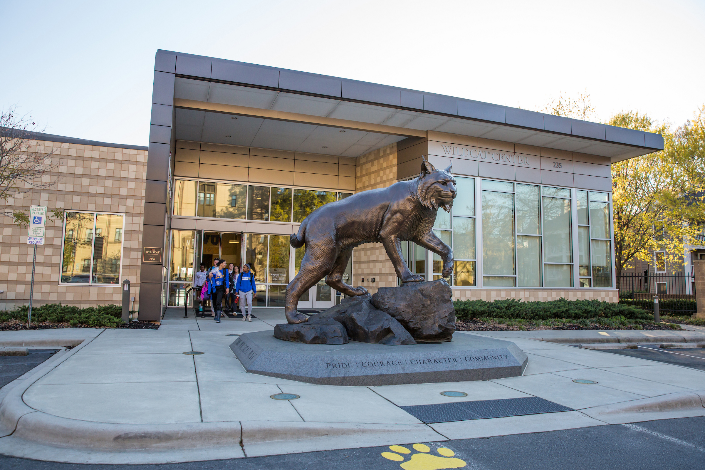A bronze statue of a wildcat stands on a pedestal in front of the Wildcat Center, with people walking toward the building's entrance.