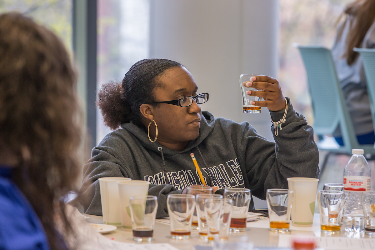 A person wearing glasses and a hoodie holds a glass of liquid while seated at a table with multiple glasses and cups, appearing to engage in a tasting or evaluation activity.