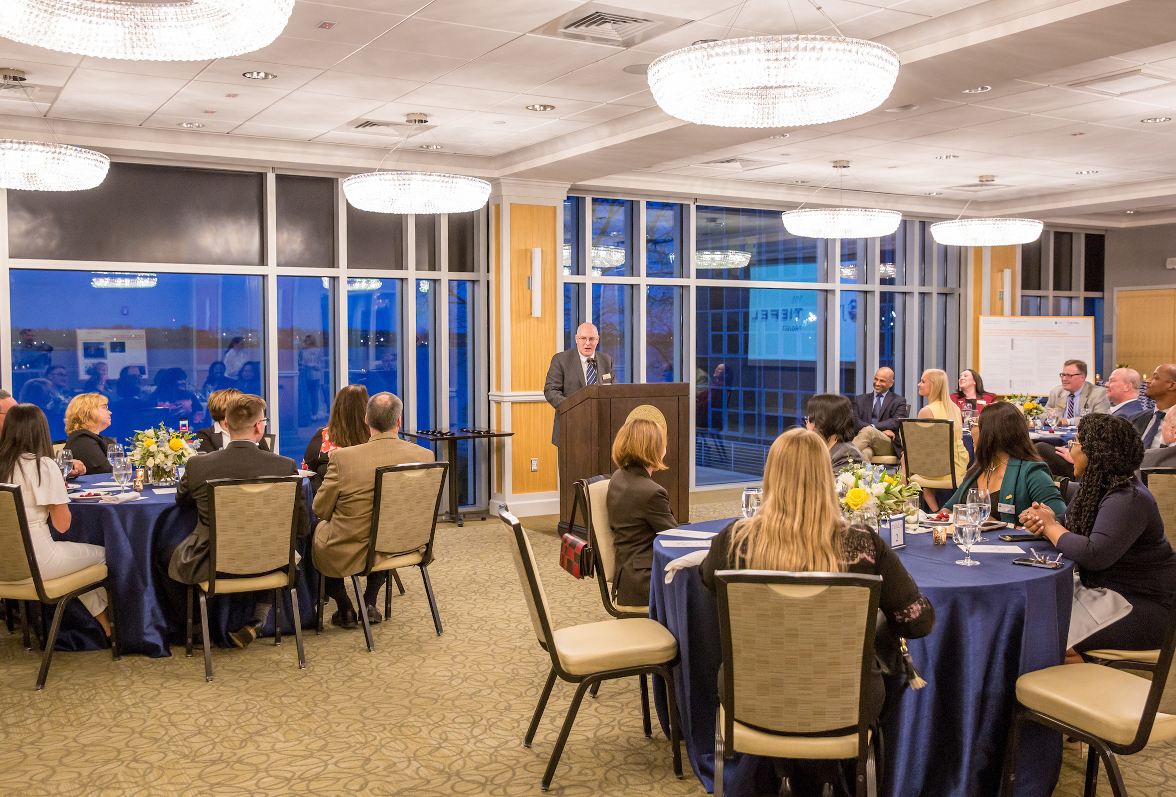 A speaker addresses a seated audience at a formal event in a well-lit banquet hall with large windows and elegant chandeliers.