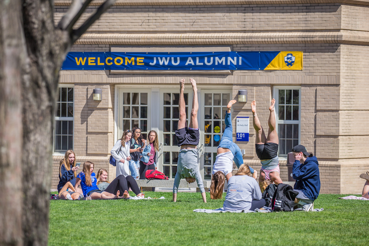 A group of people, including some performing handstands, gather on a grassy area in front of a building with a "Welcome JWU Alumni" banner.