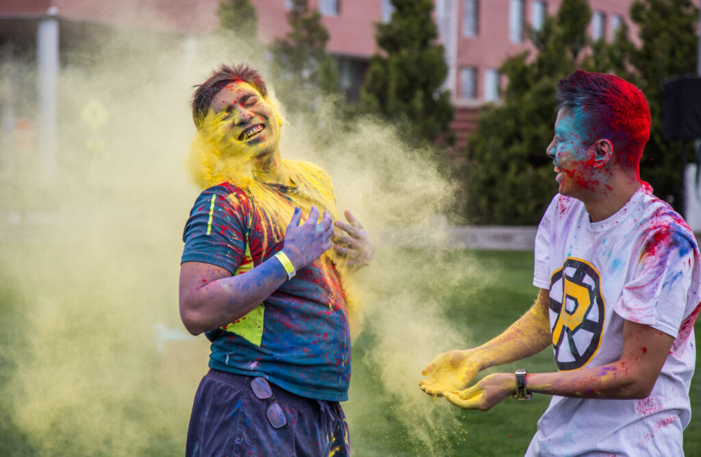 Two people covered in colorful powder smile during an outdoor campus event.