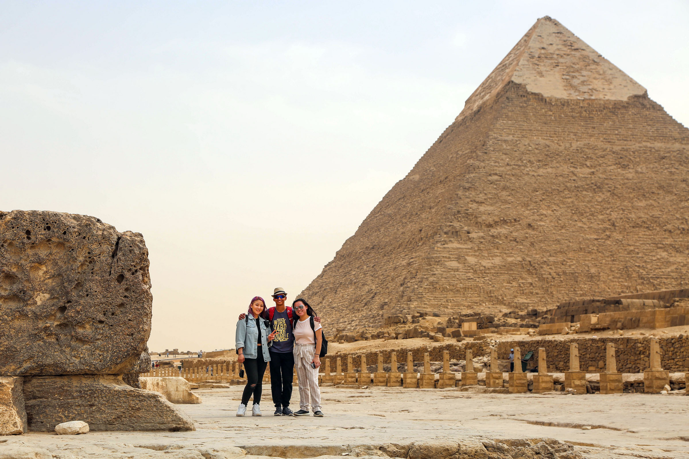 Three people pose together in front of a large pyramid in a desert setting, with ancient stone ruins visible nearby.