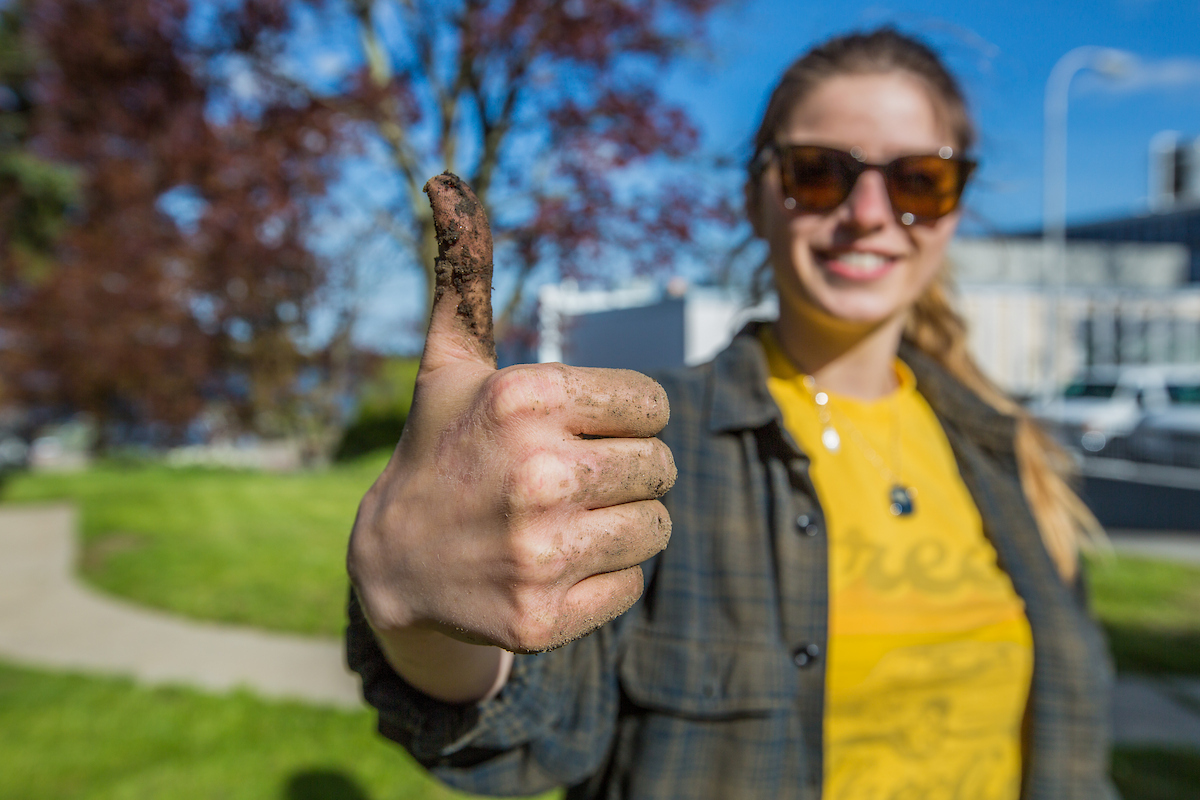 A person with sunglasses gives a thumbs-up gesture, their hand covered in dirt, against a sunny outdoor backdrop.