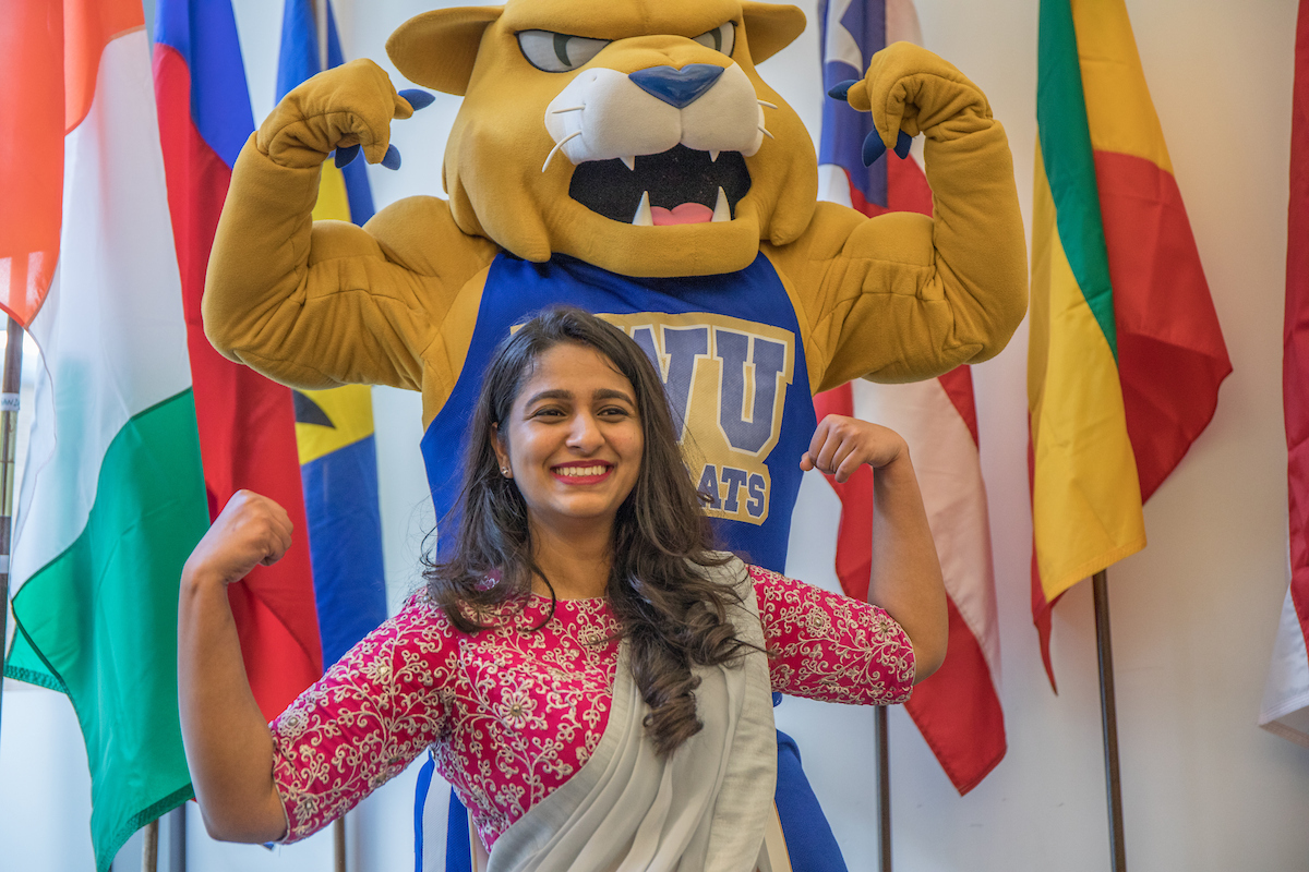 A person in a traditional garment poses with flexed arms in front of a mascot wearing a "WUATS" jersey, with international flags in the background.