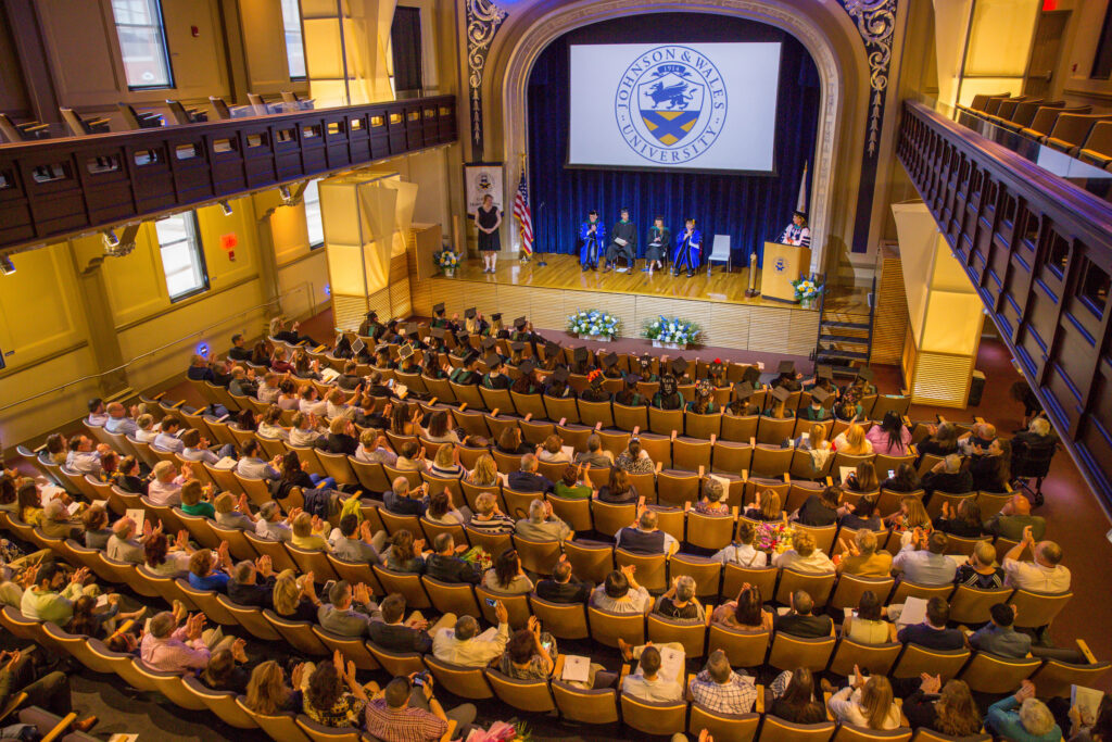 Graduates and attendees applaud during a ceremony at Johnson & Wales University, with the university's logo displayed on a screen behind the stage.