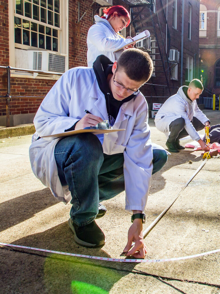 Several individuals in lab coats are conducting an outdoor measurement activity on a sidewalk between brick buildings, with bright sunlight creating lens flares.