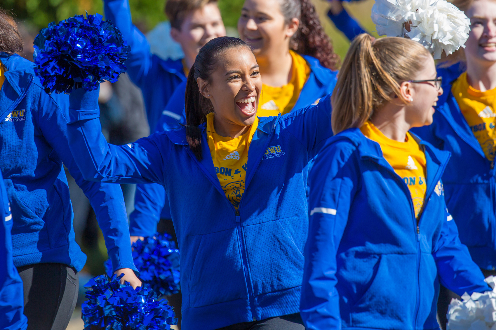 A group of cheerleaders in blue and yellow uniforms wave blue and white pom-poms while smiling during an outdoor event.