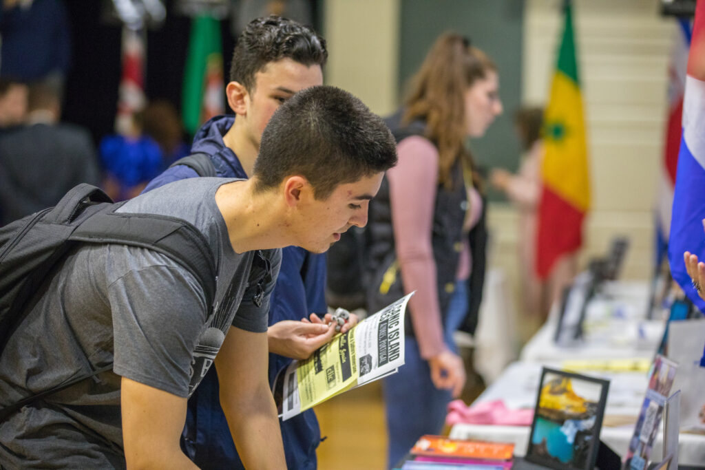 A young person with a backpack leans over a table, examining a flyer at what appears to be an international fair, with flags and other attendees visible in the background.
