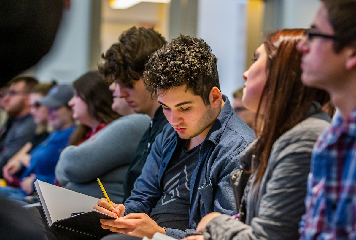 A group of individuals seated in a row, with one person in the foreground actively writing in a notebook with a pencil.
