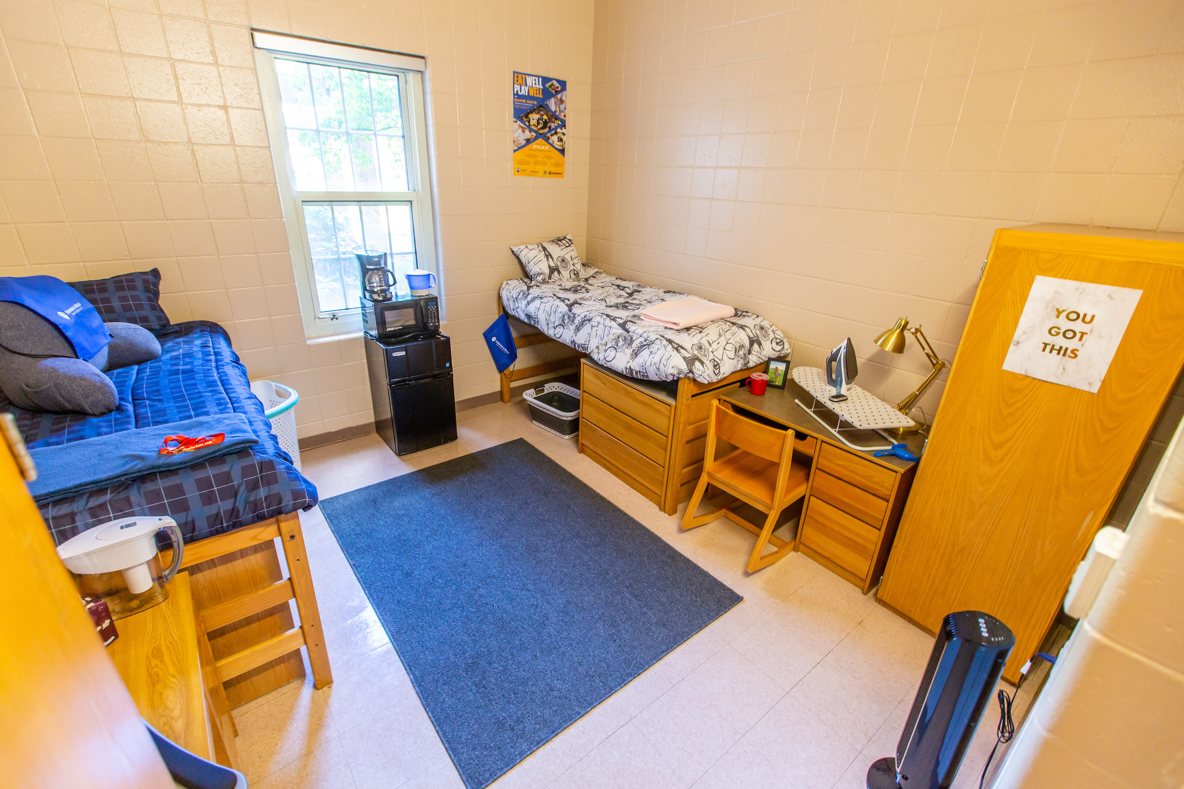 A neatly arranged dorm room with two beds, a desk, a refrigerator, and a window, featuring a motivational note on the door.