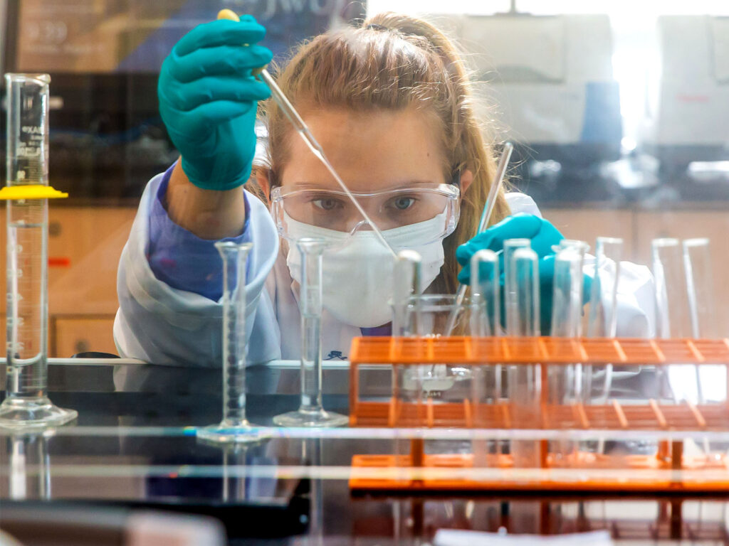 A chemistry student in a lab coat and gloves carefully transfers liquid using a pipette while wearing safety goggles and a mask.