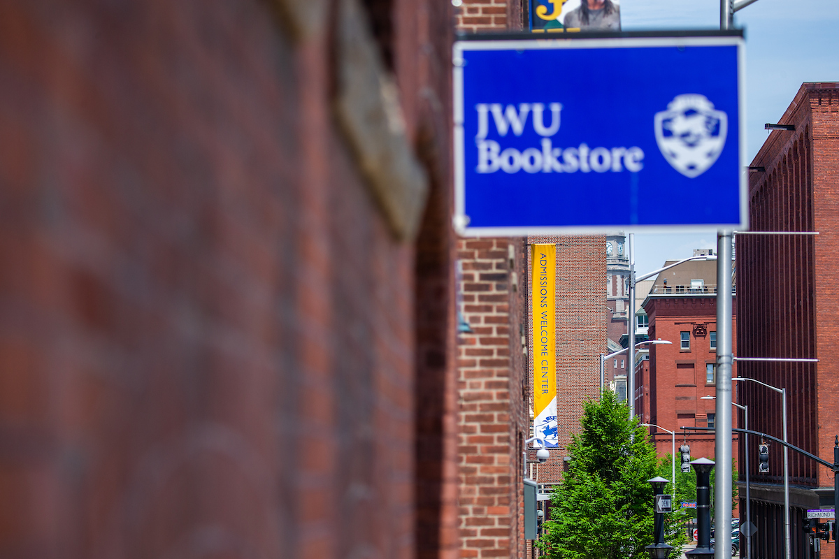 A blue sign for the JWU Bookstore hangs beside a brick building, with a yellow banner reading "Admissions Welcome Center" visible in the background.