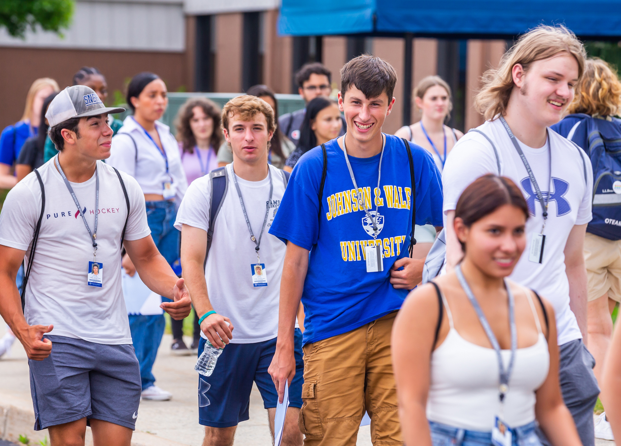 A group of young people wearing lanyards and casual attire walk together outdoors, with one person in a blue "Johnson & Wales University" shirt smiling prominently.