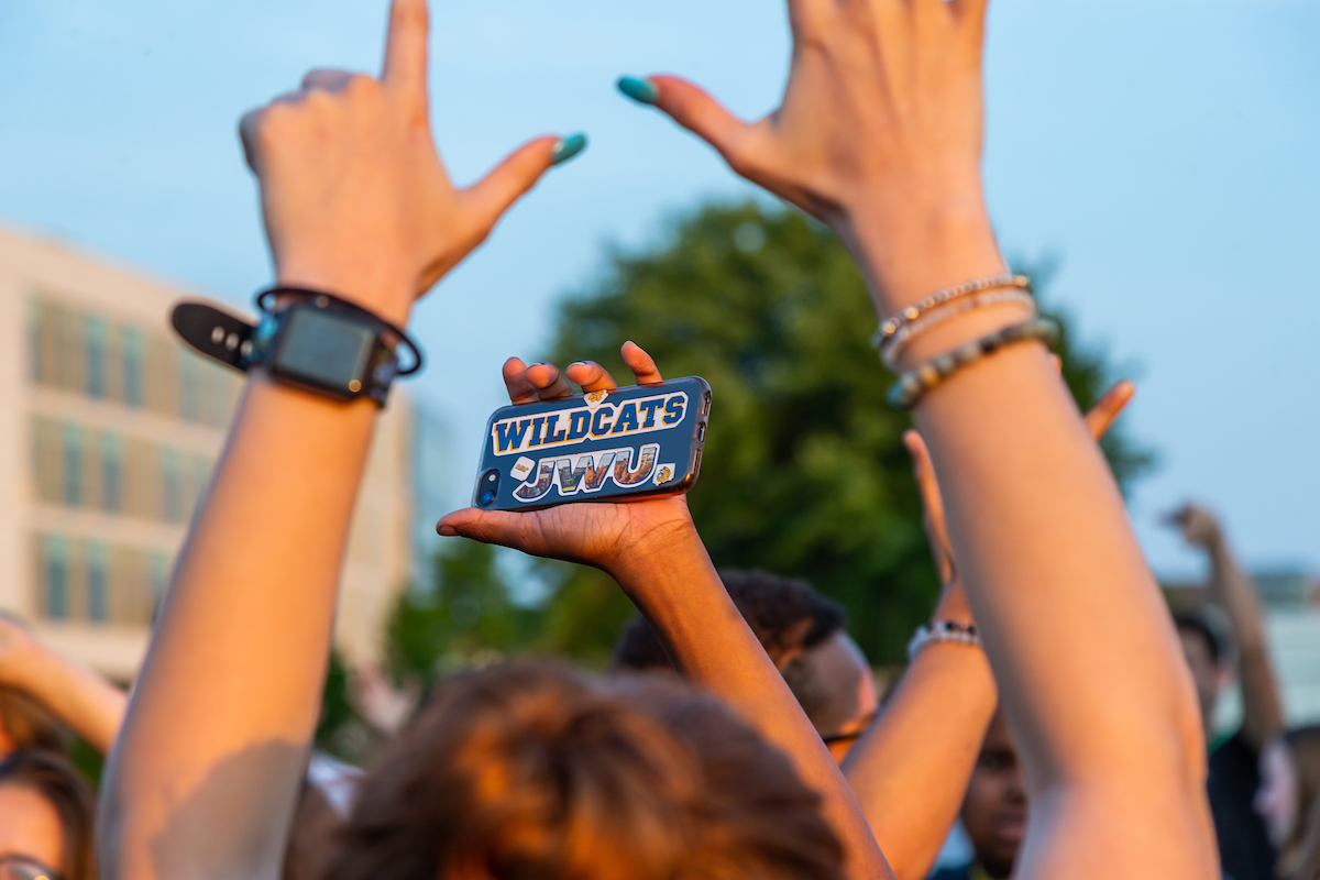 A person holds up a smartphone with a "Wildcats JWU" case while raising their hands in celebration at an outdoor event.