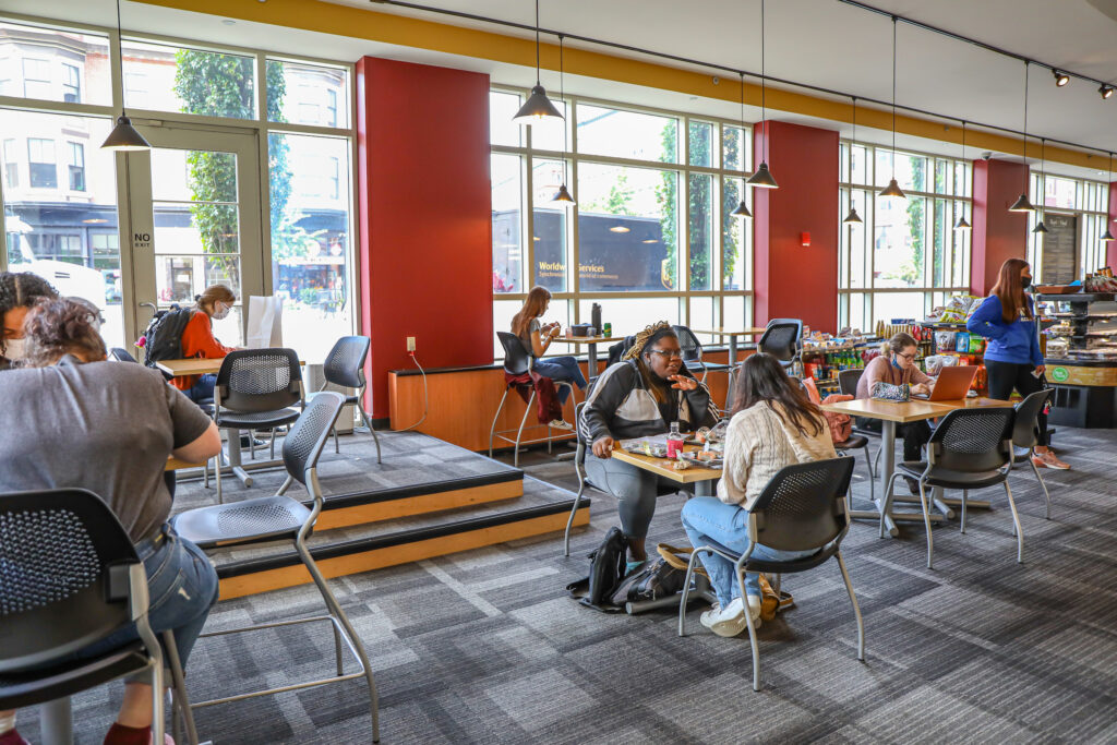 People are seated at tables in a bright, modern café with large windows and red accent walls, engaged in conversations and using laptops.