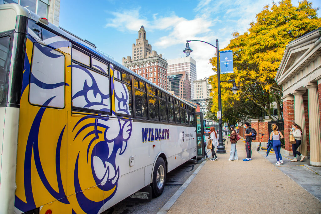 A yellow and blue Wildcats bus is parked on a city street with students walking nearby, autumn trees, and tall buildings in the background.
