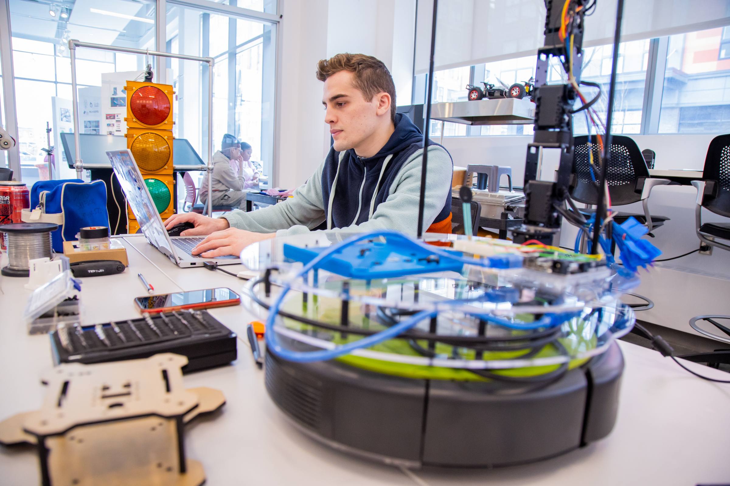 A person works on a laptop surrounded by robotics components and tools in a well-lit lab setting.