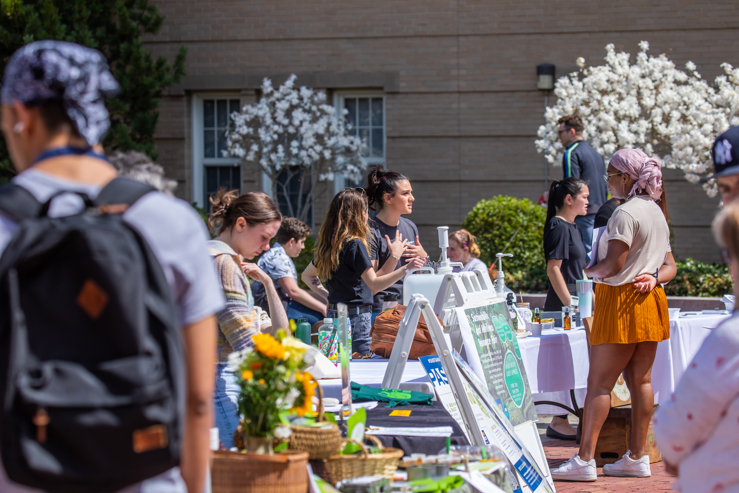 A group of people gather outdoors around tables displaying items and signs, with blooming trees and a building in the background.