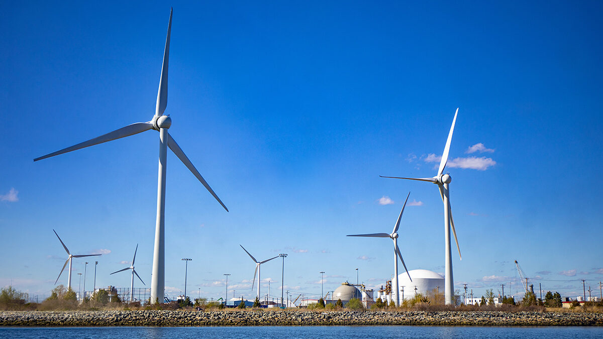 Multiple wind turbines stand along a waterfront under a clear blue sky, with industrial structures visible in the background.