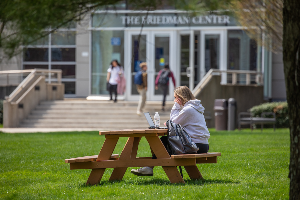 A person sits at a wooden picnic table on a grassy lawn, using a laptop, with The Friedman Center visible in the background.