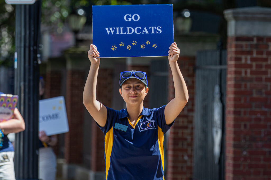 A person holds up a blue sign that reads "GO WILDCATS" with paw prints, wearing a matching team shirt and cap.