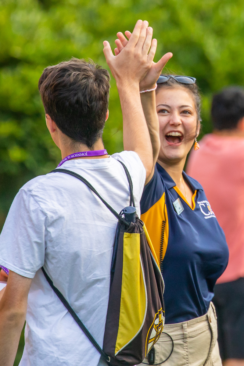 Two individuals high-five outdoors, one wearing a white shirt and a drawstring bag, the other in a blue and yellow polo shirt with sunglasses on their head, smiling joyfully.