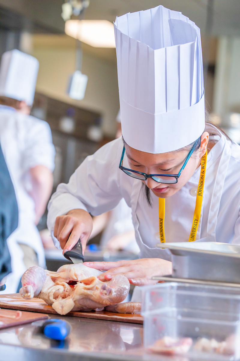 A chef in a white uniform and tall hat carefully cuts a whole chicken on a wooden board in a professional kitchen.
