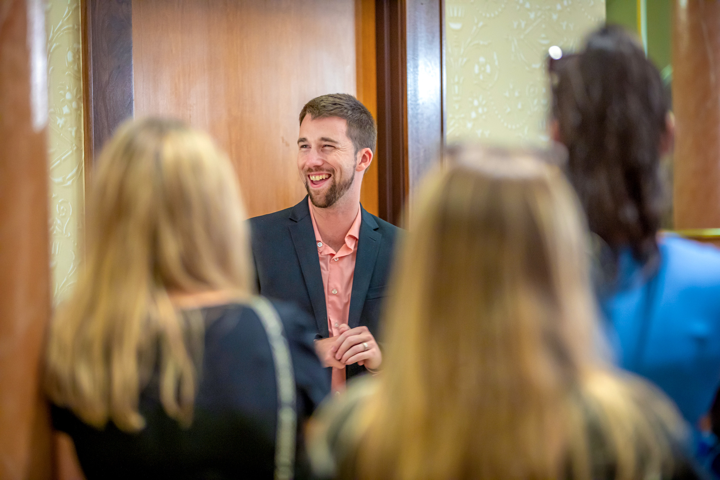A smiling person in a suit and pink shirt stands in a doorway, engaging with others in a warmly lit indoor setting.