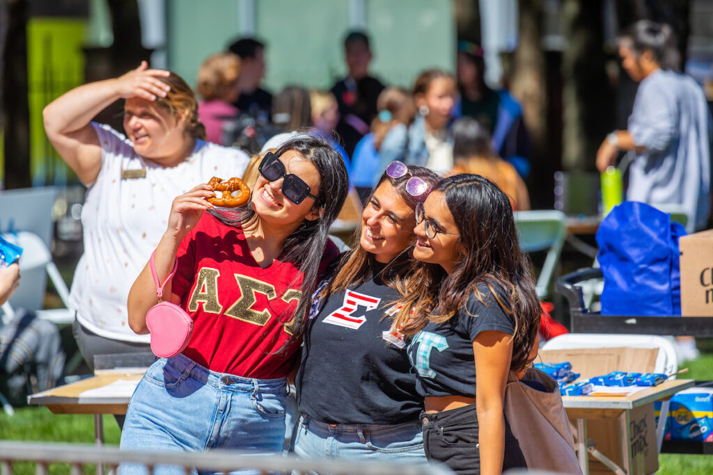 Three young people smile and pose together outdoors, one holding a pretzel, at what appears to be a campus or community event.