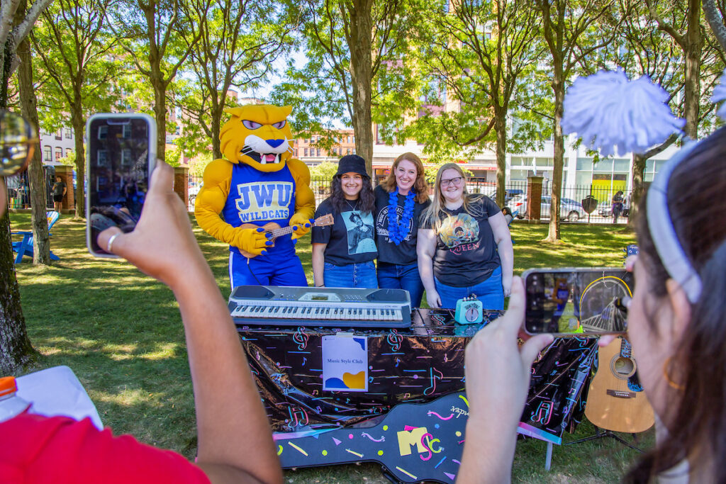 A mascot and three individuals stand behind a music-themed table outdoors, while two people photograph the scene with their phones.