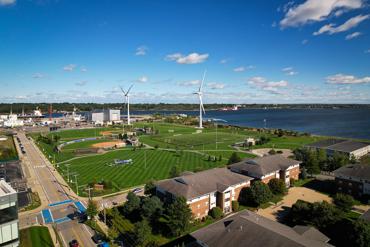 An aerial view of a waterfront area featuring wind turbines, sports fields, residential buildings, and a large body of water under a bright blue sky.