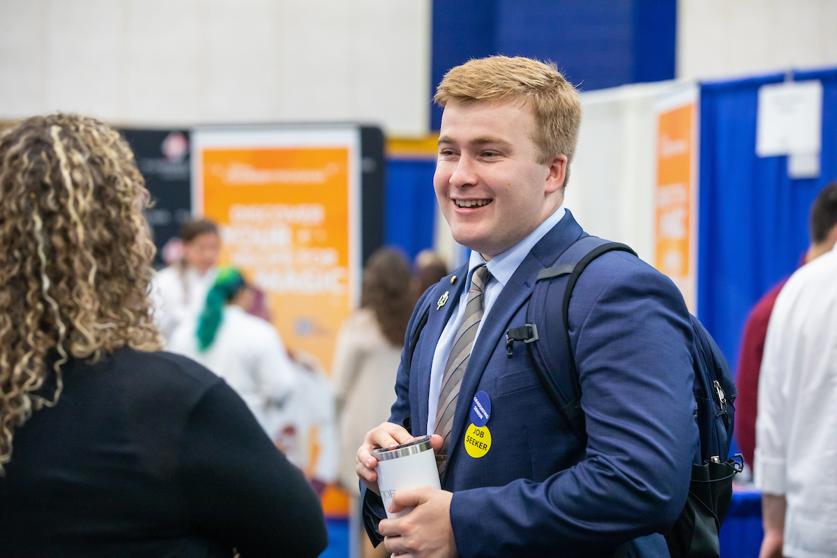 A young person in a suit and tie, wearing a "Job Seeker" badge, smiles while holding a travel mug during a career fair.