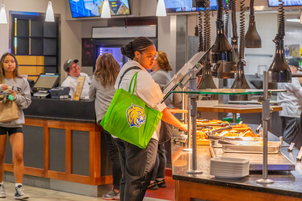 A person selects food from a buffet line in a brightly lit cafeteria, carrying a green tote bag with a mascot logo.