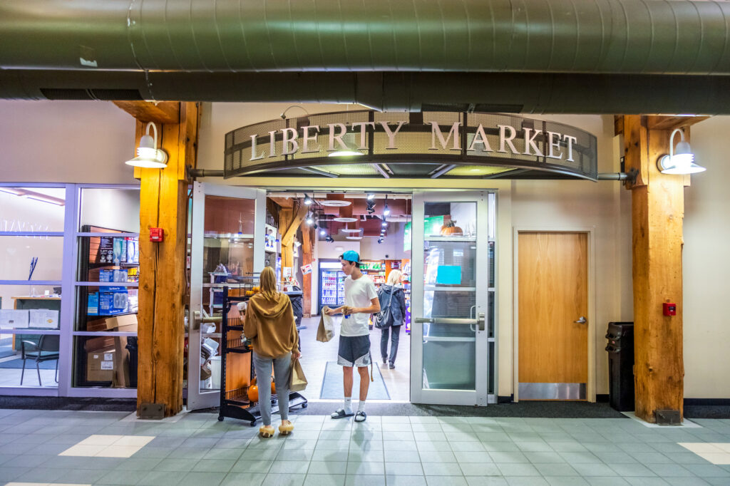 Two individuals stand at the entrance of Liberty Market, a storefront with wooden beams and glass doors, holding shopping bags.