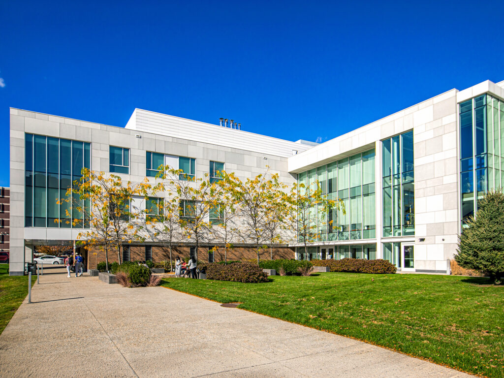 A modern, light-colored building with large glass windows and a grassy courtyard under a clear blue sky, with several people walking or standing near the entrance.