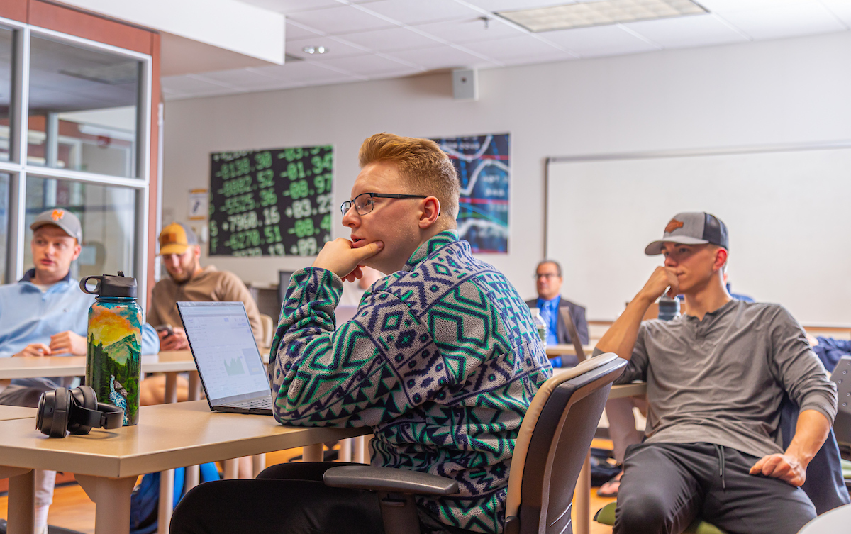 Several finance students are seated at desks in a classroom, attentively listening while one person uses a laptop and another rests their chin on their hand.