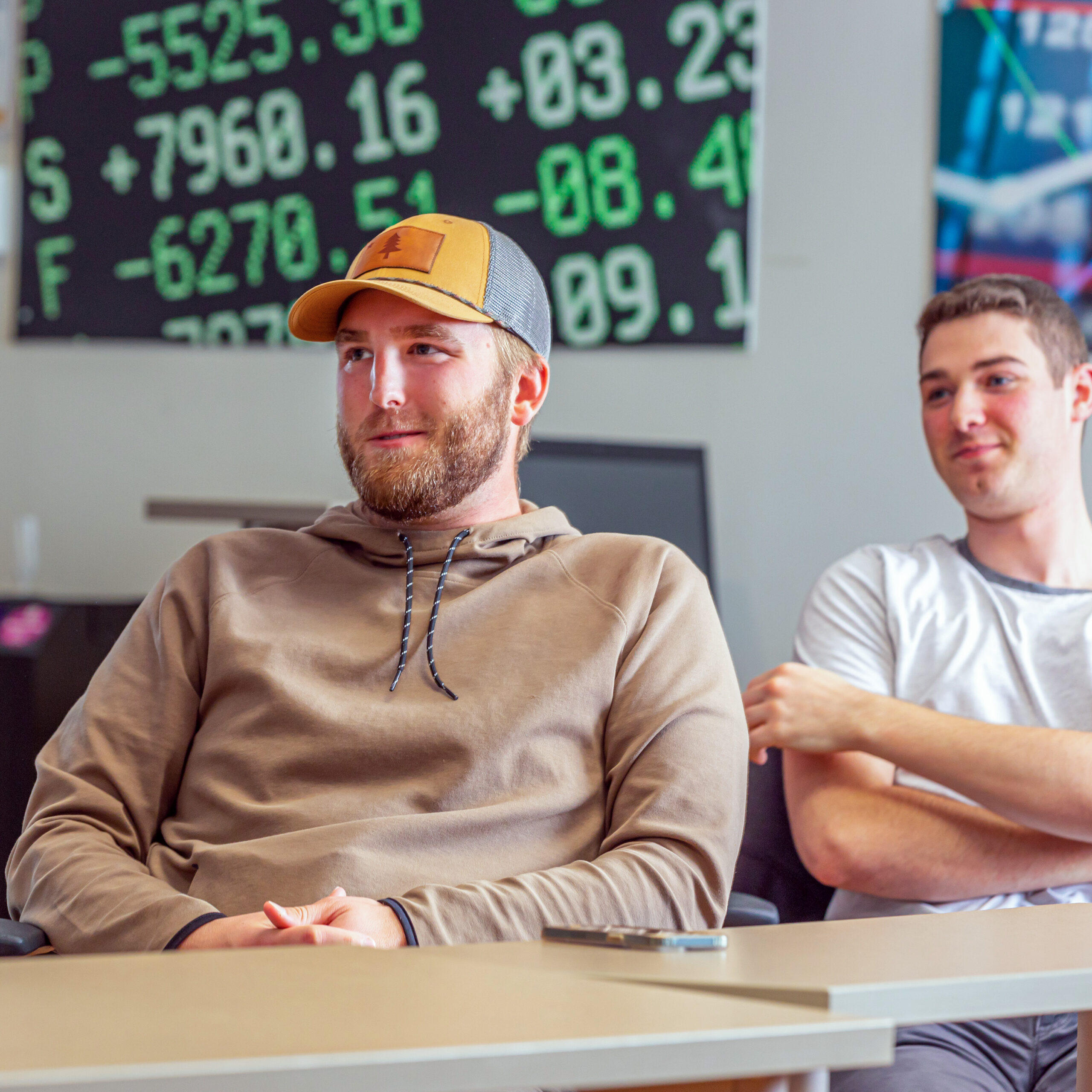 Two students sit at a table during a class discussion with a stock market display behind them.