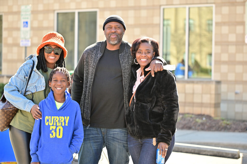 A family of four smiles for a photo outdoors, with one member wearing a blue hoodie featuring "CURRY 30" and another holding a beverage can.