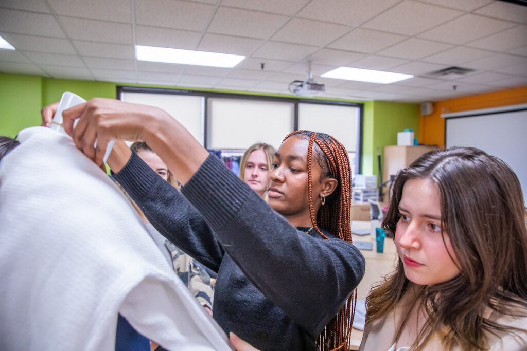 A person with braided hair holds a white garment while others observe in a classroom setting.