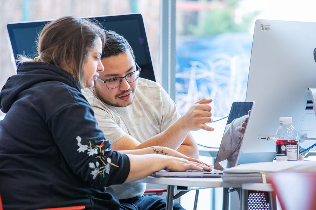 Two individuals collaborate closely at a desk, focused on a laptop screen with one gesturing toward it.